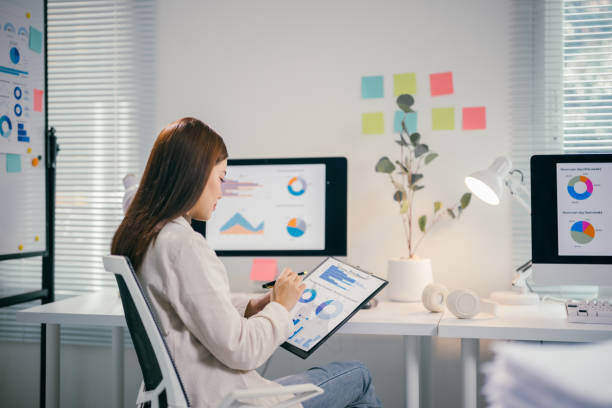 istock Young asian professional reviewing financial data on clipboard and screen, working intently at sleek corporate workspace 2195417314