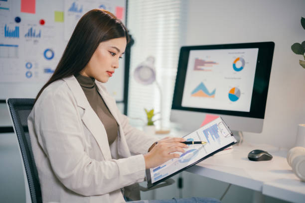 istock Professional asian female working at sleek workspace, checking financial reports on clipboard and monitoring data displayed on computer screen 2195417351