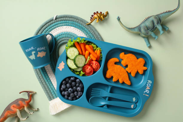 istock Dinosaur-themed kids meal set with a blue plate featuring chicken nuggets shaped like dinosaurs, fresh vegetables, blueberries, a matching cup with 'ROAR!' text, and toy dinosaurs on a woven mat 2195423667