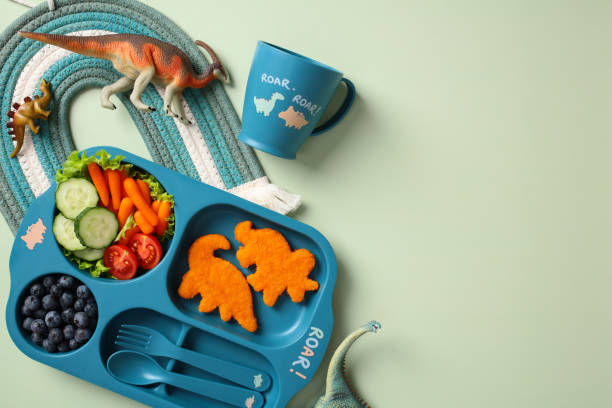istock Children's meal set with dinosaur-shaped chicken nuggets, fresh vegetables, and blueberries on a blue tray with a matching mug and toy dinosaurs on an green background 2195423849