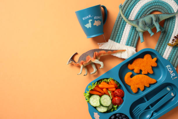 istock Dinosaur-themed kids meal set with a blue tray featuring dinosaur-shaped fish nuggets, fresh vegetables, and blueberries. Includes a matching cup, toy dinosaurs, and a woven rainbow mat 2195423906