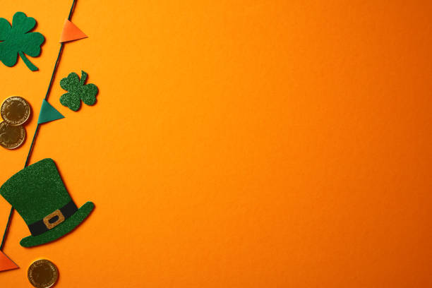 istock Happy St. Patrick's Day concept. Banner design with green glitter shamrocks, a leprechaun hat, gold coins, and colorful pennant flags on an orange backdrop 2195424331