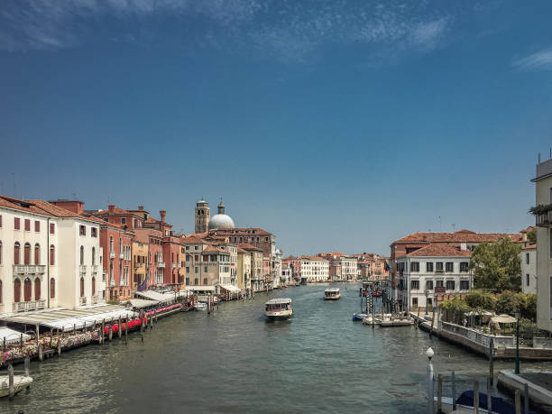 This photo captures a vibrant scene on the Grand Canal in Venice, Italy. The canal stretches into the distance, lined with colorful buildings and dotted with boats. The sky is a brilliant blue, adding to the cheerful atmosphere. The image evokes a sense of warmth and vitality, inviting the viewer to imagine a sunny day exploring Venice.