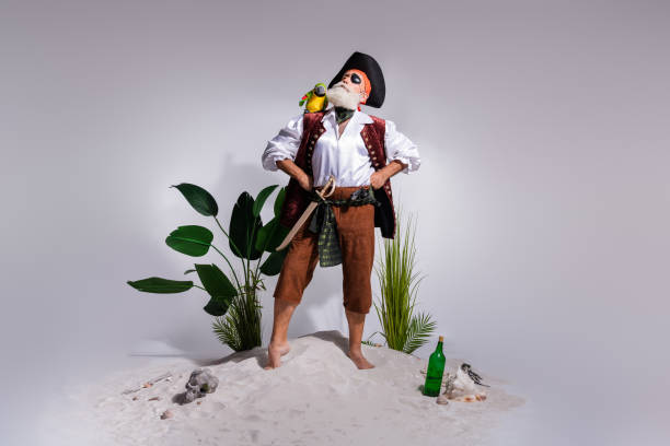 A bearded, elderly pirate stands confidently on sand, masked as a humorous character with a parrot on his shoulder. He is dressed in a buccaneer costume, surrounded by tropical plants.