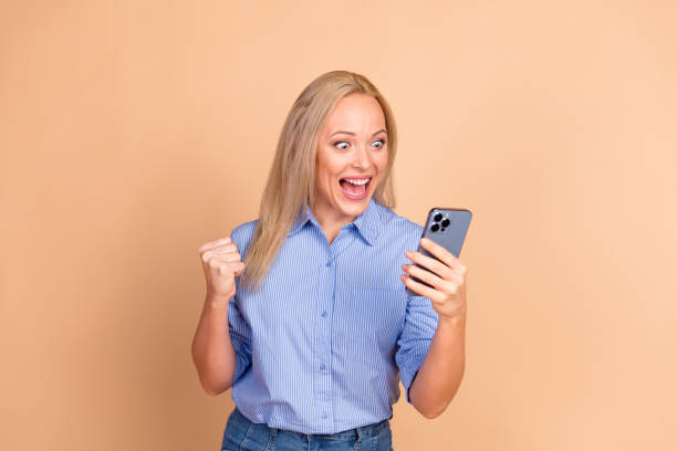 istock Excited Woman Celebrating Success While Holding Smartphone Against Beige Background in Casual Blue Striped Shirt 2195455708