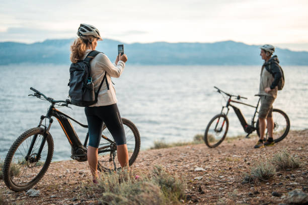 A mature Caucasian woman uses a smartphone beside her partner, both clad in casual cycling gear, with their electric mountain bikes against a coastal sunrise backdrop.