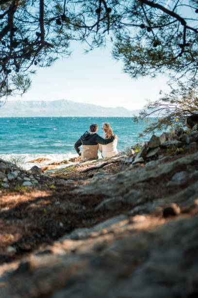 A serene mature Caucasian couple sits together enveloped by nature, gazing out at the expansive ocean vista from a tranquil beachside spot.