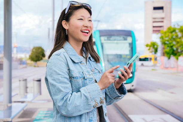 istock Smiling Young Woman Using Smartphone at Tram Stop in Urban Setting 2195463068