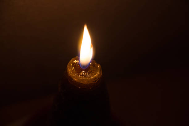 Close-up of a single lit candle. \n\n\nThe candle's flame is a bright, yellowish-orange, and appears to be burning steadily. The candle wax is a dark, rich brown color, and small details of melted wax are visible near the top. \n\n\nThe background is completely dark, providing a stark contrast to the candle's light, focusing the viewer's attention on the flame itself. The overall impression is one of warmth and subtle illumination in a dark space.