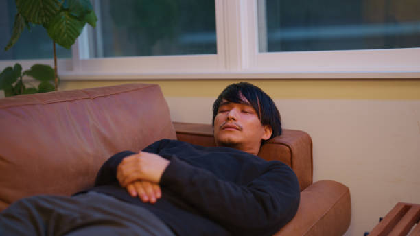 A young man lies on a couch with his hands folded over his chest, appearing relaxed during a mental health therapy session. The peaceful setting with soft lighting and a modern interior fosters a sense of calm, providing a supportive space for emotional healing and introspection.