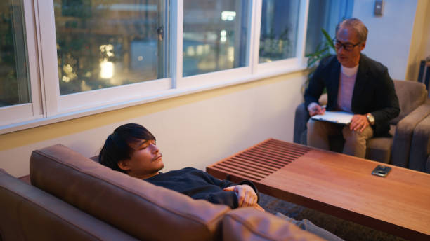 A young man lies on a couch during a mental health therapy session, engaging in conversation with his therapist, who listens attentively while taking notes.