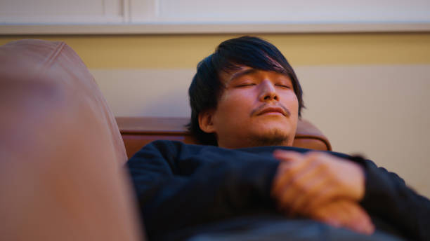 A young man lies on a couch with his hands folded over his chest, appearing relaxed during a mental health therapy session. The peaceful setting with soft lighting and a modern interior fosters a sense of calm, providing a supportive space for emotional healing and introspection.
