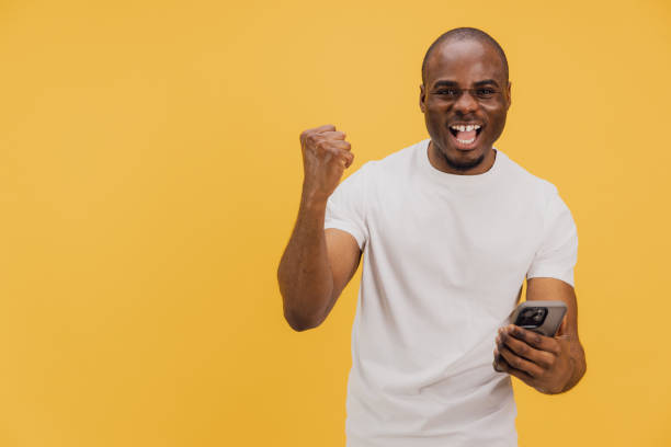 istock Excited man celebrating success while holding smartphone on yellow background 2195477263
