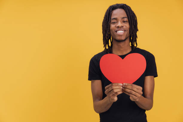 istock Young man holding a big red heart, smiling on yellow background 2195480430