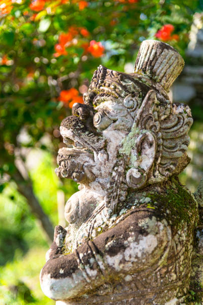 A beautifully crafted Balinese stone statue stands beside colorful tropical flowers, showcasing intricate details and cultural artistry, illuminated by sunlight in a lush outdoor environment.