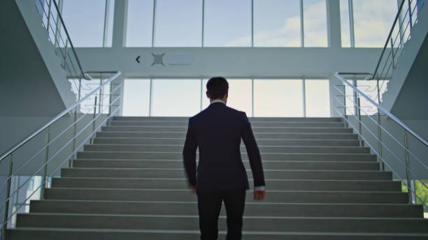 istock Back view businessman climbing modern glass stairway in bright office interior. 2195492596