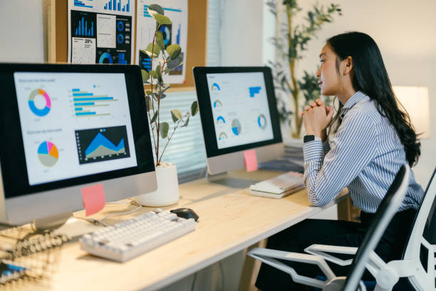 istock Young businesswoman sitting at desk analyzing financial data displayed on computer screens, working late in modern office with charts and graphs on wall 2195503415