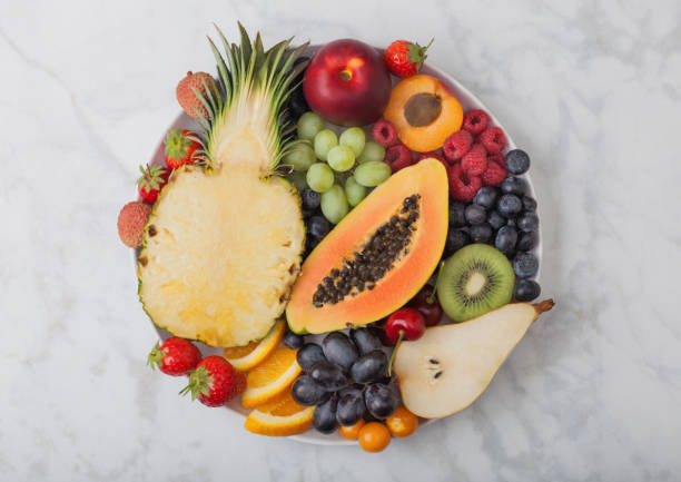 Fresh raw organic summer berries and exotic fruits in white plate on marble background. Pineapple, papaya, grapes, nectarine, orange, apricot, kiwi, pear, lychees, cherry and physalis. stock photo