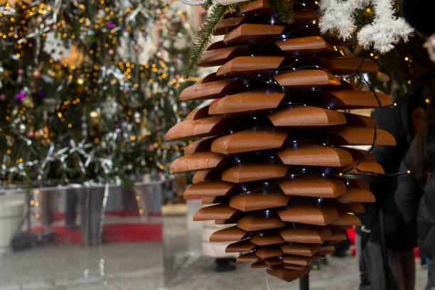 A large pinecone-shaped decoration constructed from wooden slats is illuminated by soft lights, adding a warm touch to a festive holiday display surrounded by trees.