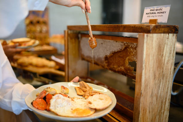 A person adds natural honey to a delicious breakfast spread featuring eggs, toast, and vegetables. This inviting morning scene highlights a unique honeycomb dispenser, enhancing the fresh and wholesome dining experience.