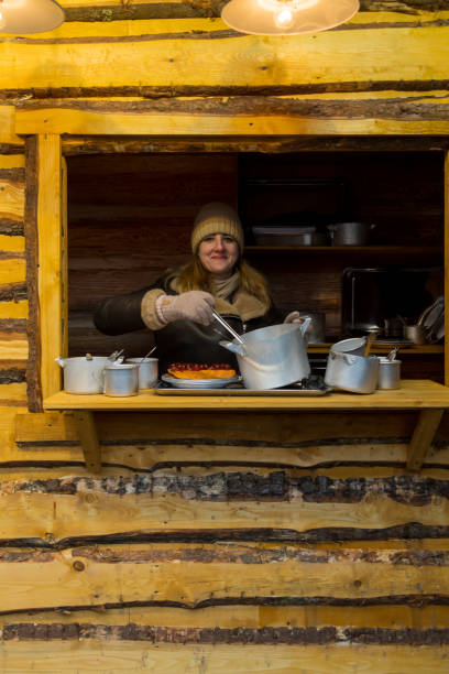 In a warm wooden cabin, a woman prepares and serves delicious hot food. The setting is wintery and mountainous, creating a cozy atmosphere for visitors.