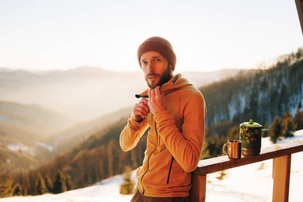 A portrait of a handsome man dressed in a cozy, warm fleece jacket, standing amidst the serene winter mountains during sunset