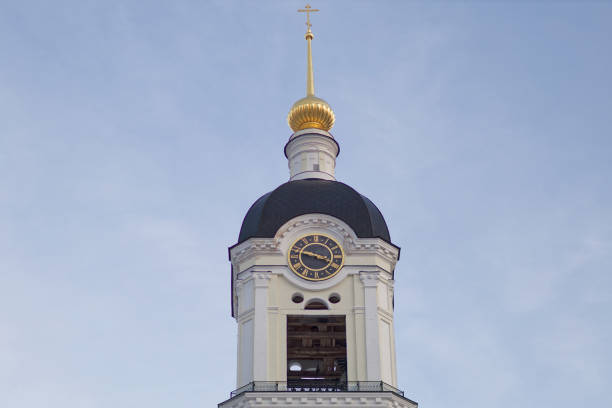 A white church tower in a small European city from the side with a visible clock and a metal roof with a Christian cross on top of it. The clock shows 6 o'clock time.
