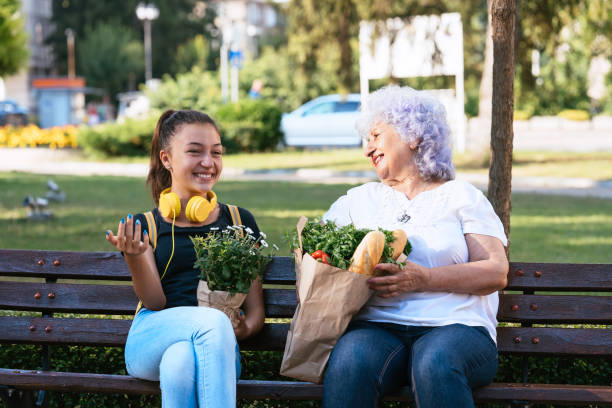 Grandma and teen enjoy nature on park bench chatting A young woman with headphones laughs with an elderly lady holding groceries on a park bench, capturing a moment of joy and bonding across generations. teenage girl with grandmother stock pictures, royalty-free photos & images