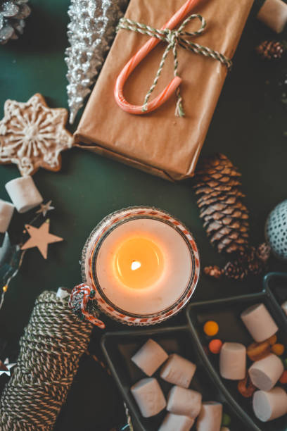 istock Cozy Christmas setup featuring a wrapped gift with a candy cane, a lit candle, tea, an open book, pinecones, marshmallows, and evergreen branches on a green background 2195619709