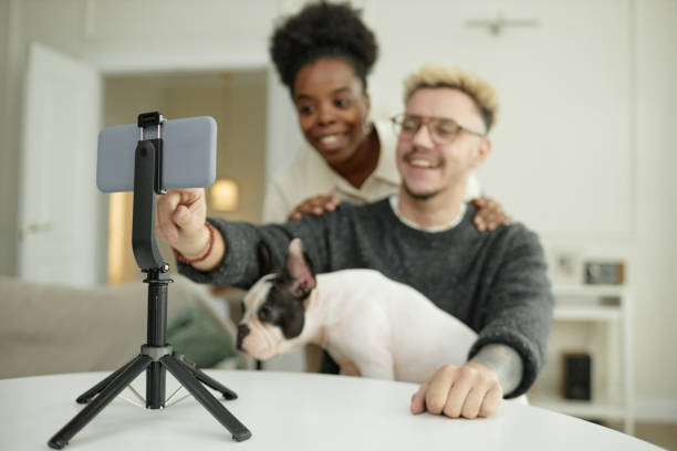 istock Happy Multiethnic Couple Recording Video Using Phone Mounted on Tripod at Home 2195628829