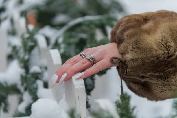 A hand, elegantly decorated with multiple rings, gently rests on a white fence blanketed in snow. The scene captures the contrast of winter elements and natural beauty.