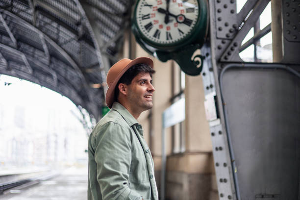 Young tourist wearing a stylish hat waits at the train station under a large vintage clock, smiling while gazing off into the distance, filled with anticipation for the journey ahead