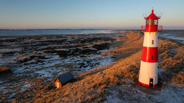 Red and white striped lighthouse situated on a coastal landscape at sunrise in winter. Lighthouse West-List on the Ellenbogen (the \