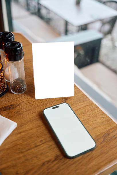 Blank vertical menu and smartphone with white screen placed on wooden table near condiments in contemporary cafe