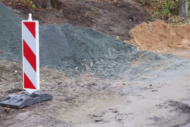 A road, on the side of which a red and white barrier is installed, behind which construction material is piled up.