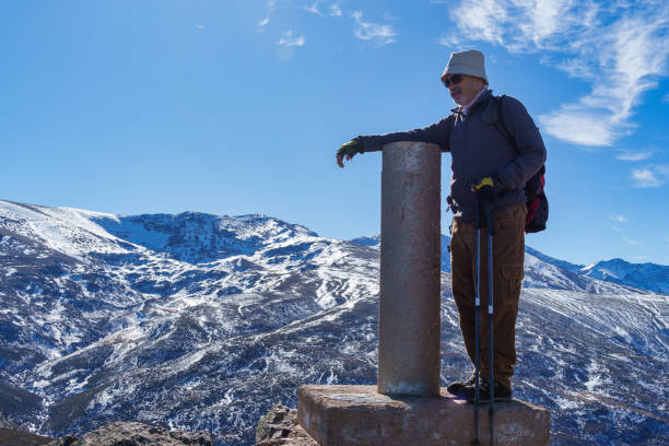 A senior man leans on a summit marker with trekking poles in Sierra Nevada, Granada, enjoying the snowy mountain views under a clear blue sky.