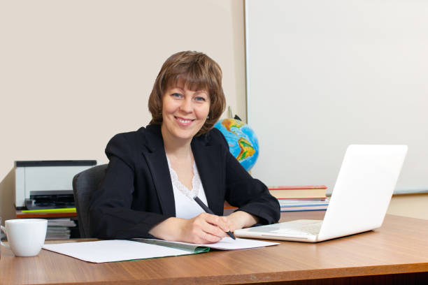 A young pretty teacher sits at her desk in the classroom and smiles at the camera. There is a laptop on the desk. There is a world map hanging on the wall behind her. A friendly teacher in a school classroom.