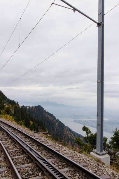 Train tracks on a mountainside with stunning lake and mountain views in the distance.  A cloudy day.