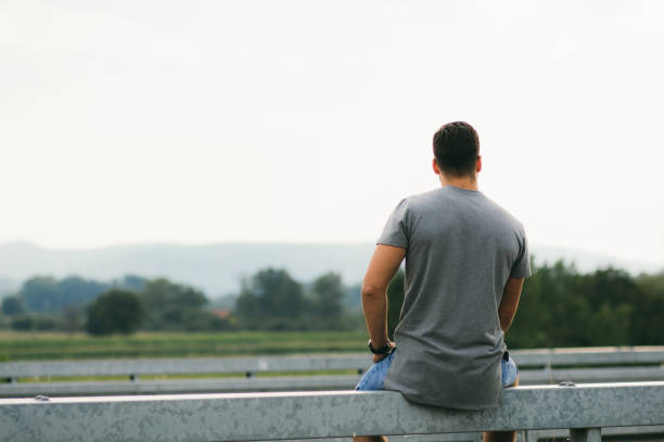 A man sits on a railing outdoors holding a longboard, contemplating amidst scenic landscapes in a calm atmosphere, representing outdoor leisure, lifestyle, and personal reflection in a serene setting