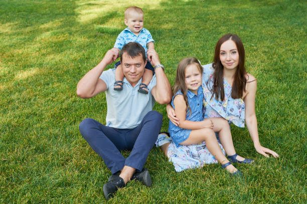 A small family of four sit together on a blanket in a park as they enjoy a picnic together.  They are each dressed casually and are smiling as they enjoy spending time together.