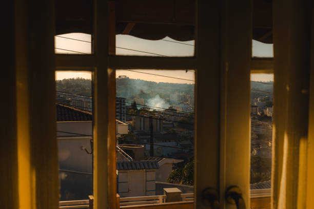 A view of a cityscape at sunset, framed by a window and illuminated by warm golden-hour light. In the distance, smoke rises from a visible fire, adding a dramatic element to the serene urban scene. The contrast between the cozy interior perspective and the unfolding event outside creates a striking composition.