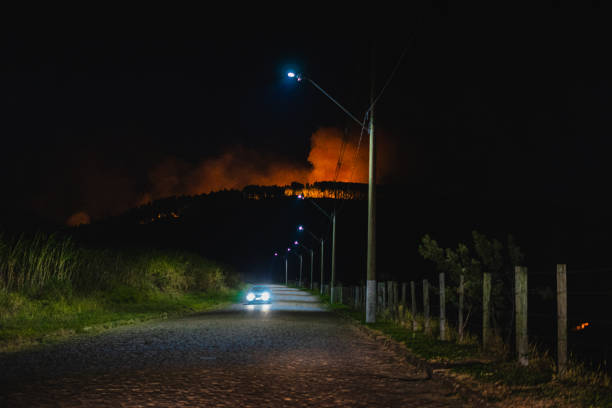 A rural cobblestone road illuminated by streetlights and a car’s headlights leads toward a distant forest fire lighting up the night sky. The glowing flames engulf the tree line on a hillside, contrasting with the dark surroundings. The scene captures the tension between human infrastructure and the natural disaster unfolding in the background.