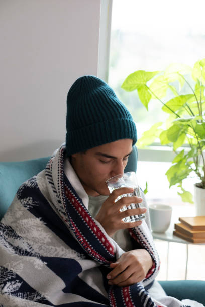 istock Sick young man drinking water wrapped in blanket at home 2195754205
