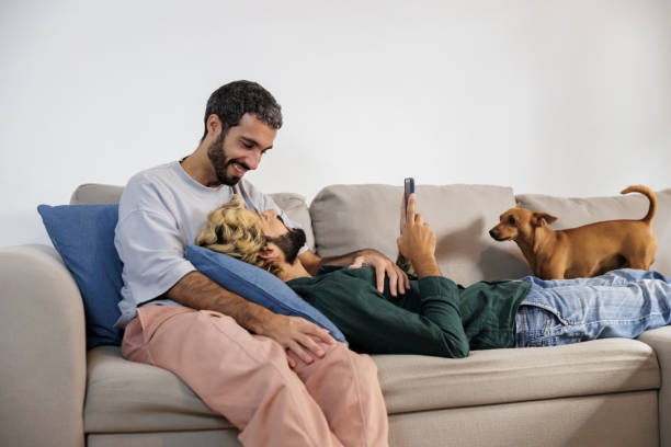 istock Gay couple relaxing on sofa with smartphone and dog 2195766577