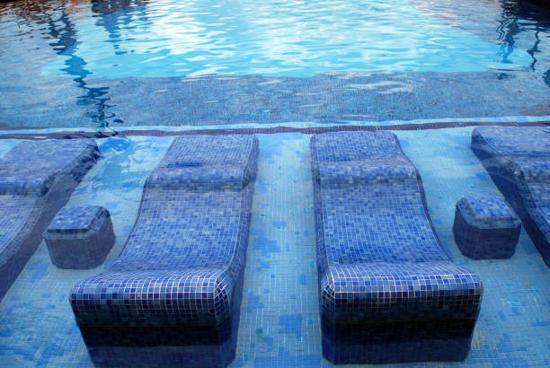 Subject: Horizontal parital view from the back of a sunbathing woman sitting on a beach lounge chair. She wears a red swimsuit and an aqua blue, straw sun hat with a big, floppy brim. She has a view of a pristine swimming pool, palapas, and the blue sky over the Caribbean Sea. The aqua blue sea water matches the color of her hat.