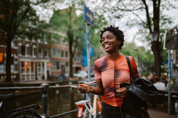 Amsterdam street style fashion. Portrait of a stylish black young woman with short hair on the beautiful streets of Amsterdam, wearing an orange shirt and a jacket. She is walking the streets, checking apps and messages on her smartphone.