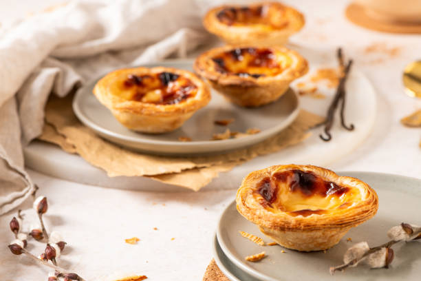 Several Pastel de Nata are arranged on plates with cinnamon, vanilla beans, and small golden spoons.
