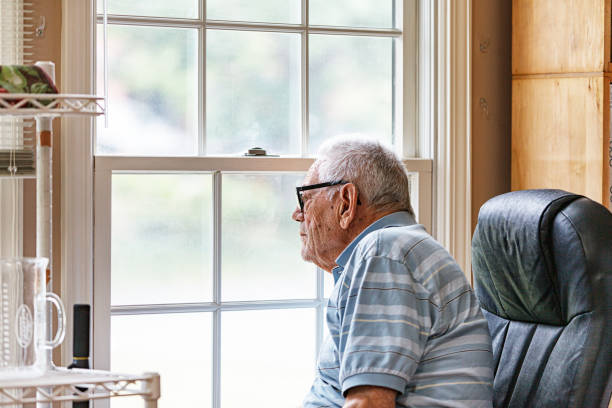 A real life, real person, hearing impaired elderly senior adult man wearing a hearing aid is sitting up intently as he looks outside curiously through the hazy, screened living room window at home. He has early stage dementia and often sits alone observing the world outside his window.