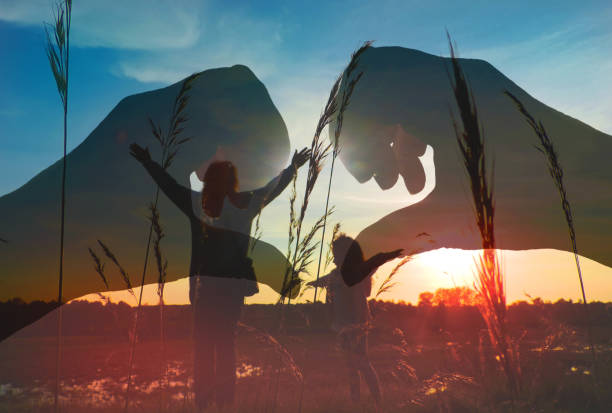 Multiple exposure, silhouette of hands making a heart shape finger frame, Idyllic sunset landscape and child with mother in the meadow with raised hands, rear view. Love, hope, pray. Enjoying Life