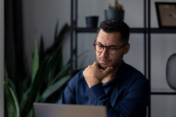 Pensive businessman working from home using laptop and thinking with hand on chin